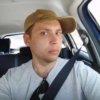 Selfie of a man in a beige cap and gray T-shirt sitting in a car with the seatbelt on